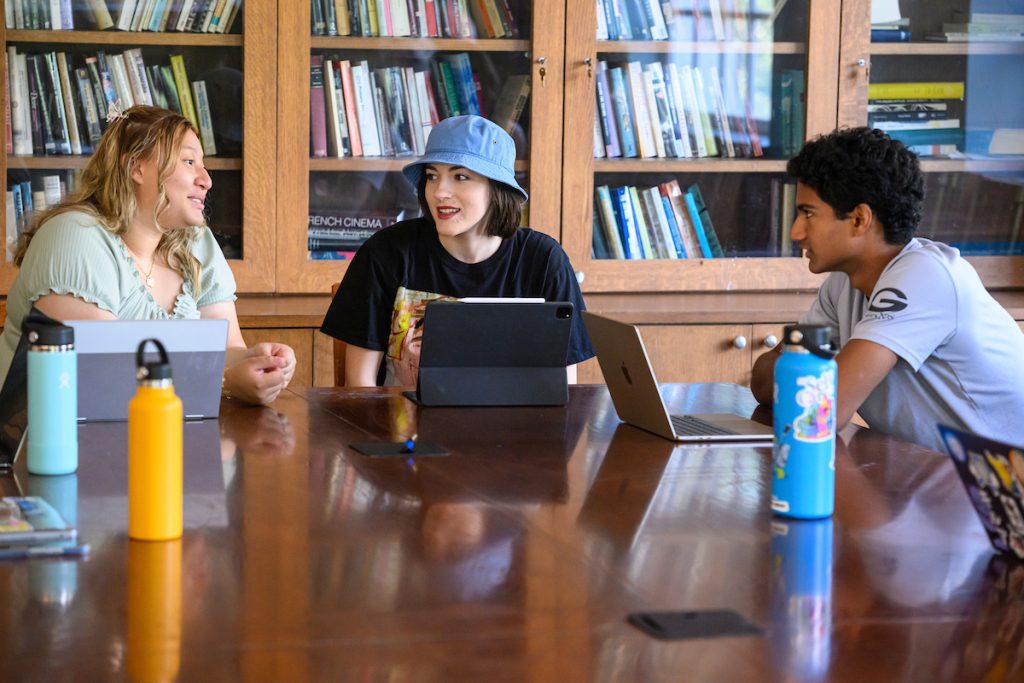 Three students sitting at a table with a book, talking together